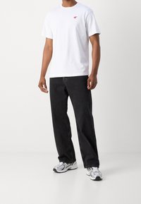 Weißes T-Shirt mit kurzen Ärmeln und kleinem roten Logo auf der Brust, kombiniert mit schwarzen Jeans und grauen Sportschuhen. Schlichter, lässiger Look.