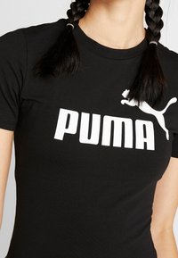 Czarna koszulka z krótkim rękawem z białym logo Puma, noszona przez osobę z dwoma splecionymi warkoczami widocznymi na ramionach.