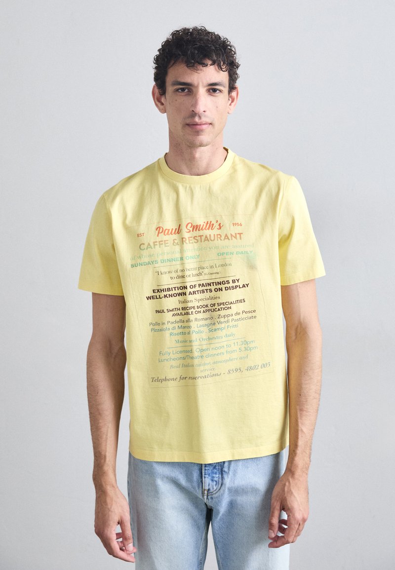 Yellow cotton T-shirt featuring printed restaurant text in various colors. Short sleeves, crew neck, relaxed fit, and casual denim jeans.