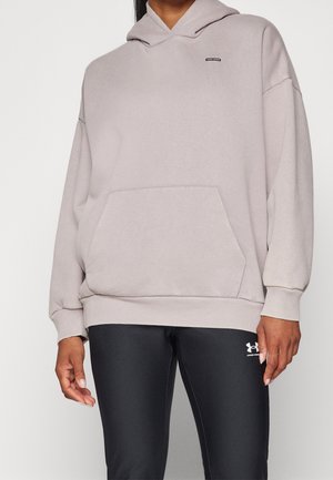 Persoon draagt een lichtgrijze oversized hoodie met kangoeroezak en zwarte Under Armour legging, staand tegen een effen achtergrond.