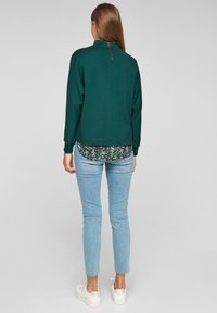 Türkiser Pullover mit hohem Kragen und Reißverschluss, über einem blumigen Hemd getragen. Kombiniert mit hellblauen, engen Jeans und weißen Sneakers.