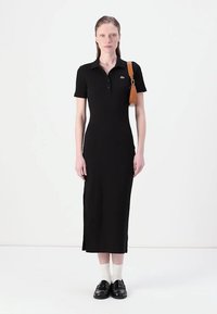 Juoda polo suknelė trumpomis rankovėmis, prigludusio silueto ir vidutinio ilgio. Yra šoninė įplyšimo detalė ir nedidelis logotipas ant krūtinės. Suderinta su juodais loafer'iais.