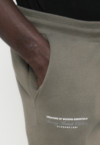 Calças de moletão verde-azeitona com um cós canelado e bolsos laterais. Apresenta texto branco na parte da frente: "CRIADORES DE ESSENCIAIS MODERNOS."