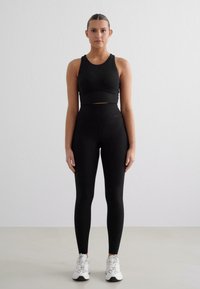 Melna sportiska crop top un augsta vidukļa legingi, pieguļošs, elastīgs audums, minimālistisks dizains. Baltie sporta apavi noslēdz apģērbu.