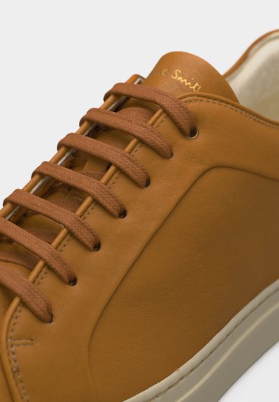 Κοντινό πλάνο καφέ δερμάτινου αθλητικού παπουτσιού με κορδόνια ίδιου χρώματος και μπεζ σόλα, που δείχνει λεπτομερή ραφή και το όνομα της μάρκας στη γλώσσα.