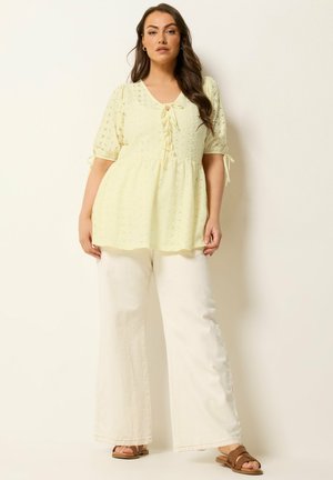 Kvinde iført en bleg gul bluse med hulmønster, bindebånd og pufærmer, hvide bukser med vidde i benene og brune sandaler, stående foran en ensfarvet baggrund.