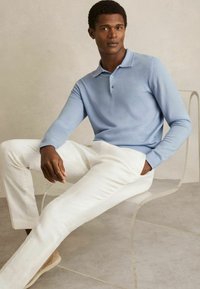 Hellblaues Langarm-Polo aus weichem Stoff, mit einer Zwei-Knopf-Leiste. Kombiniert mit weißen Leinenhosen und beigen Schuhen.