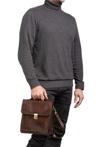 Cartella in pelle marrone con patta, accessori in oro e cinturino staccabile. Tenuta da una persona che indossa un maglione grigio a collo alto e jeans neri.