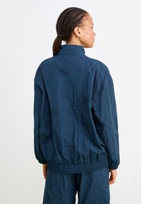 Tumši zils Nike vēja žakets ar elastīgām aprocēm un apakšmalu, ar izteiksmīgu zaļu izšūtu logo uz muguras un augstu apkakles dizainu.