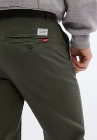 Άτομο που φοράει παντελόνι Levi's σε ελιά πράσινο χρώμα με πίσω τσέπη και εμφανή ετικέτα με το λογότυπο Levi's, ντυμένο με ανοιχτό γκρι φούτερ.