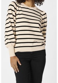 Nainen, jolla on päällä vaalean kermanvärinen neule mustilla vaakaraitoja ja mustat housut, seisoo ja yksi käsi lepää hänen reidellään.