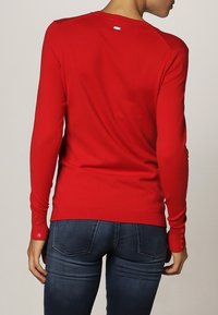 Sweater rouge à manches longues au design ajusté, avec des poignets côtelés agrémentés de détails en boutons et un petit logo près de l'encolure. Porté avec un jean foncé.
