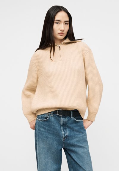 Jeune femme aux longs cheveux noirs portant un pull beige côtelé à demi-fermeture éclair et un jean bleu taille haute avec une ceinture noire.