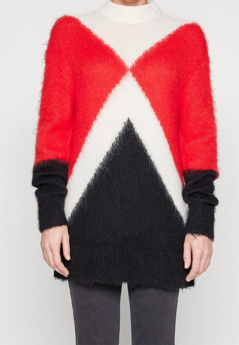 Pull en maille douce avec motif chevron géométrique rouge et noir, manches longues et col montant, porté avec un pantalon foncé.