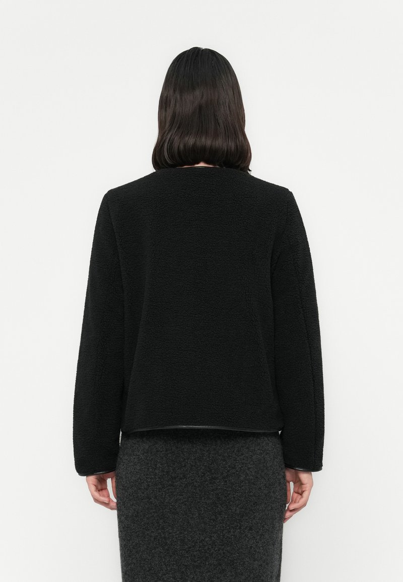 Personne avec des cheveux bruns mi-longs portant une veste noire texturée et une jupe gris foncé, se tenant face à un mur blanc.