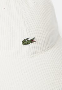 Cappello di corduroy bianco con logo di coccodrillo ricamato in verde sulla parte anteriore. Il materiale ha un aspetto testurizzato e rigato.