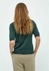 Top verde de punto con mangas cortas y puños acanalados. Textura suave, diseño ligeramente ajustado, combinado con pantalones beiges de corte ajustado.