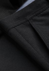 Tissu noir avec un motif fin et texturé, comportant des coutures et des poches ajustées. Doublure intérieure lisse visible le long des bords.