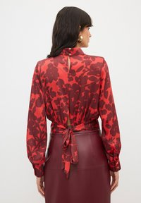 Rode bloemenblouse met lange mouwen, strikdetail op de rug en een lichte keyhole bij de hals, gecombineerd met een bordeauxleren rok. Glanzende textuur.
