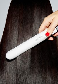 Hand met rode nagellak die een witte ghd chronos haarstylers gebruikt op lang, donker, glanzend, zijdezacht haar.