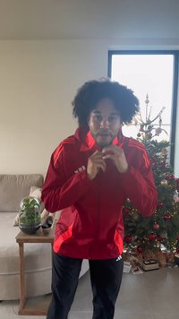 Giacca sportiva rossa con cappuccio e cerniere, con strisce bianche e nere sulle spalle. Indossata con pantaloni neri, in un ambiente interno decorato.