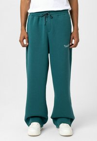 Teal joggings gemaakt van zachte stof, met een trekkoord in de taille, zijzakken en een logo op de linker bovenbeen. G gedragen met witte sneakers.