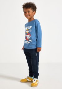 Blaues Langarmhemd mit Paw Patrol-Grafik, kombiniert mit marineblauen Jogginghosen mit strukturierten Akzenten und gelben Sneakern mit weißen Details.