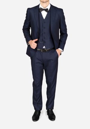 Homme portant un costume trois pièces bleu foncé avec un nœud papillon noir, une chemise blanche, une ceinture noire et des chaussures habillées noires, debout avec une main dans la poche.