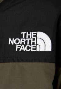 Melna un olīvu zaļa auduma ar teksturētu apdari; satur balto izšūto "The North Face" logo ar izliekta dizaina akcentiem.