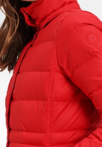 Röd puffjacka med hög krage, med vadderade sektioner och ett runt logotypmärke på axeln. Slät, glänsande tygstruktur.