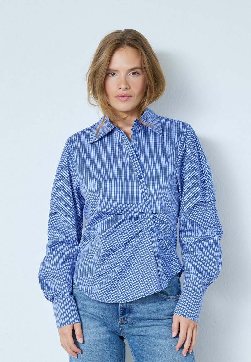 Sinivalkoraidallinen napillinen paita, jossa on terävä kaulus ja ryppyiset yksityiskohdat, yhdistetty vaaleansinisiin farkkuihin. Rentosärmä, pitkät hihat.