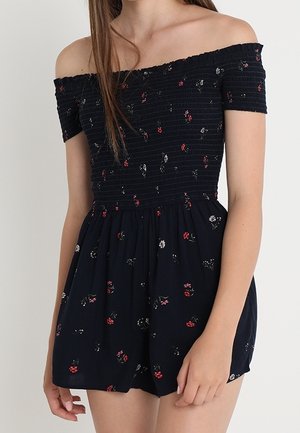 Nainen pukeutuneena mustaan olkapäät paljastavaan haalariin, jossa on pieniä punaisia ja valkoisia kukkakuvioita, röyhelöity yläosa ja lyhyet hihat.