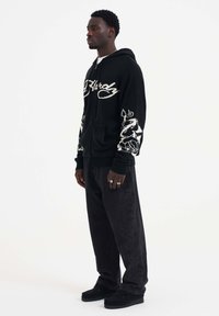 Sweat à capuche zippé noir avec broderie florale blanche sur les manches, associé à un pantalon sombre et des chaussures noires. Comprend une poche avant et des poignets côtelés.