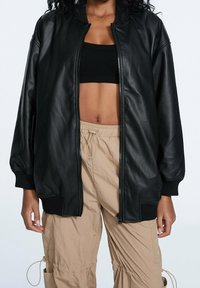 Blouson aviateur en cuir synthétique noir avec poignets et ourlet côtelés, doté d'une fermeture éclair intégrale et d'épaules tombantes. Porté sur un crop top noir.
