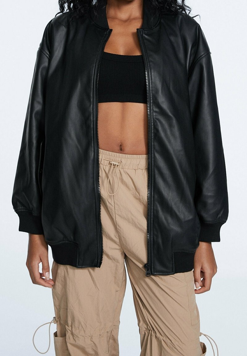 Blouson aviateur en cuir synthétique noir avec poignets et ourlet côtelés, doté d'une fermeture éclair intégrale et d'épaules tombantes. Porté sur un crop top noir.