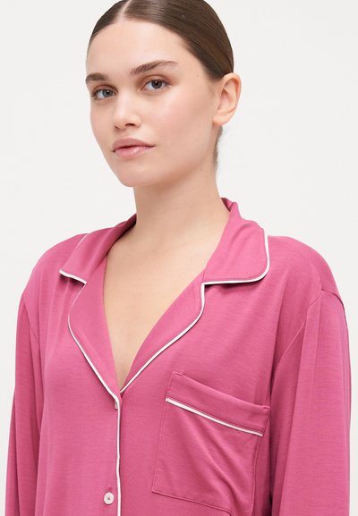 Ružová pyžamová súprava s výstrihom do V, kontrastným bielym lemovaním a náprsníkou. Mäkka tkanina s voľným strihom.