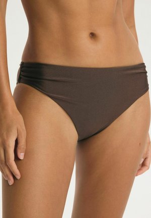 Frau trägt dunkelbraune Bikinihöschen mit seitlichen Falten, zeigt den Mittelteil und die oberen Oberschenkel vor einem einfarbigen Hintergrund.