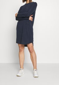 Dunkelblaues langärmliges Kleid mit rundem Halsausschnitt und gerafftem Taillenbereich, kombiniert mit weißen Sneakers und vor einem schlichten Hintergrund stehend.
