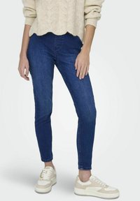 Jeans ajustados en denim azul oscuro, con una cintura de altura media. Combinados con zapatillas crema con acentos beige y cordones blancos. Enfoque en el ajuste de las piernas.