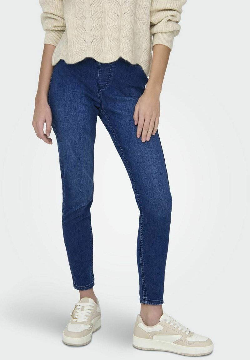 Jeans ajustados en denim azul oscuro, con una cintura de altura media. Combinados con zapatillas crema con acentos beige y cordones blancos. Enfoque en el ajuste de las piernas.