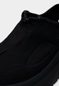 Μαύρο παπούτσι τύπου sneaker χωρίς κορδόνια με πάνελ από δίχτυ, ελαστικό κούμπωμα με τακούνι και μαλακή σόλα, σε κοντινή λήψη που δείχνει λεπτομέρειες από το επάνω μέρος μπροστά και πλάι.