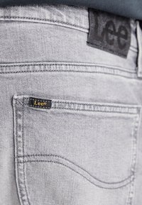 Jeans de mezclilla gris claro con una etiqueta de tela gris oscuro y costuras amarillas. Presentan un bolsillo trasero con una etiqueta negra que dice "Lee."