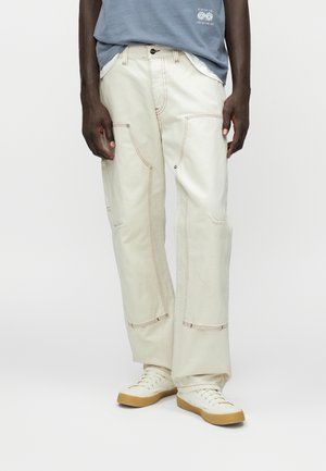 Beige cargobukser lavet af bomuld med synlige syninger og flere lommer. Bruges med en lyseblå t-shirt og hvide sneakers.