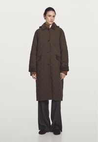 Kviltet brun jakke med hætte, store knapper og sidelommer; lagdelte brune stofdetaljer ved manchetter og krave.