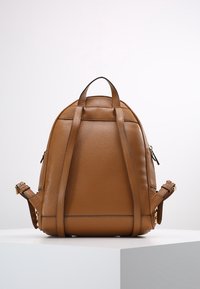 Ruskea nahkareppu, jossa on säädettävät olkahihnat ja kantokahva, esitelty valkoisella pöllyllä yksinkertaista taustaa vasten.