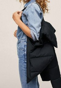 Sort polstret jakke med en glat tekstur, der har en høj krave og sidelommer, båret over en lyseblå denimskjorte og lyse jeans.