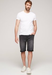 Camiseta blanca de cuello redondo y pantalones cortos de mezclilla oscuros con dobladillos enrollados. El modelo lleva zapatillas blancas; el atuendo es casual y ajustado. Fondo neutro.