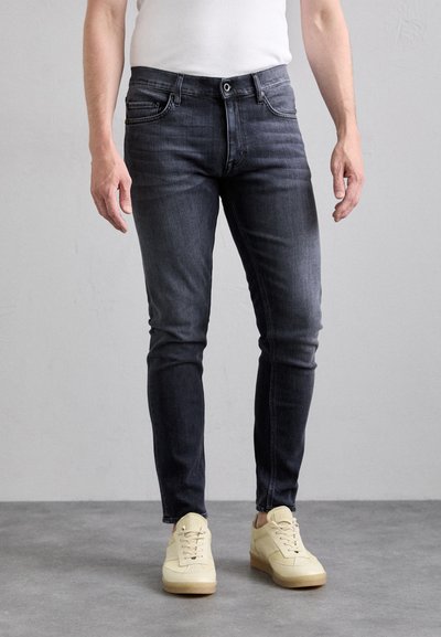 Slim-fit tmavé džíny s pěti kapsami, jemným vyblednutím a klasickým knoflíkovým zapínáním, v kombinaci se světlými teniskami.