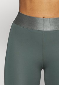 Leggings met hoge taille in donkergroene stof met een gladde textuur, voorzien van een contrasterende metalen tailleband en flatlocknaden.