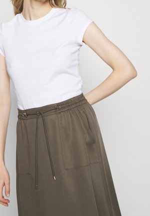 Žena nosící jednoduché bílé tričko s krátkým rukávem zastrčené do hnědé sukně s vysokým pasem, předními kapsami a šňůrkou v pase.